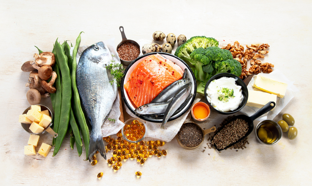 Produkty bogate w witaminę D rozłożone na białym tle — łosoś, ryba dorada, sardynki, jajka przepiórcze, brokuły, grzyby, fasola, orzechy, masło, jogurt, nasiona chia, oliwa i kapsułki suplementu — ilustracja do artykułu o suplementacji witaminy D3
